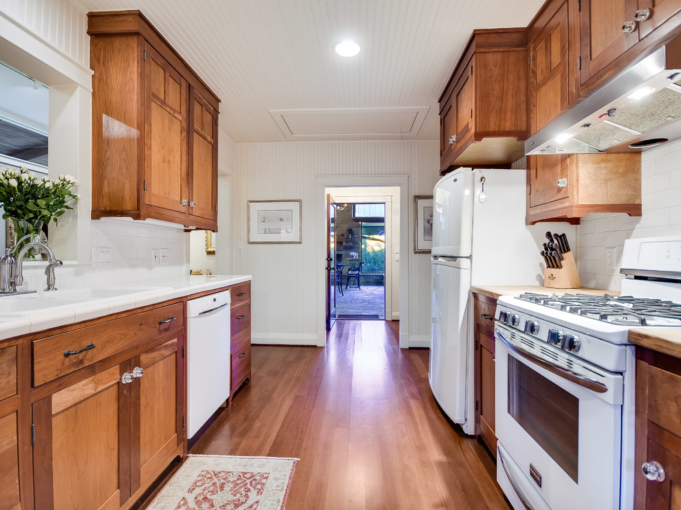 Inspiration for a medium sized classic galley kitchen pantry in Austin with a built-in sink, recessed-panel cabinets, medium wood cabinets, tile countertops, white splashback, metro tiled splashback, white appliances, medium hardwood flooring and no island.