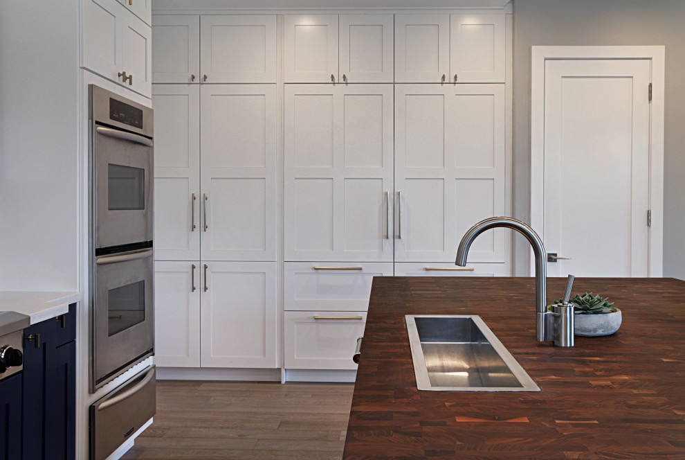 Foto på ett stort funkis brun kök, med en nedsänkt diskho, skåp i shakerstil, vita skåp, träbänkskiva, vitt stänkskydd, stänkskydd i sten, integrerade vitvaror, ljust trägolv, en köksö och grått golv