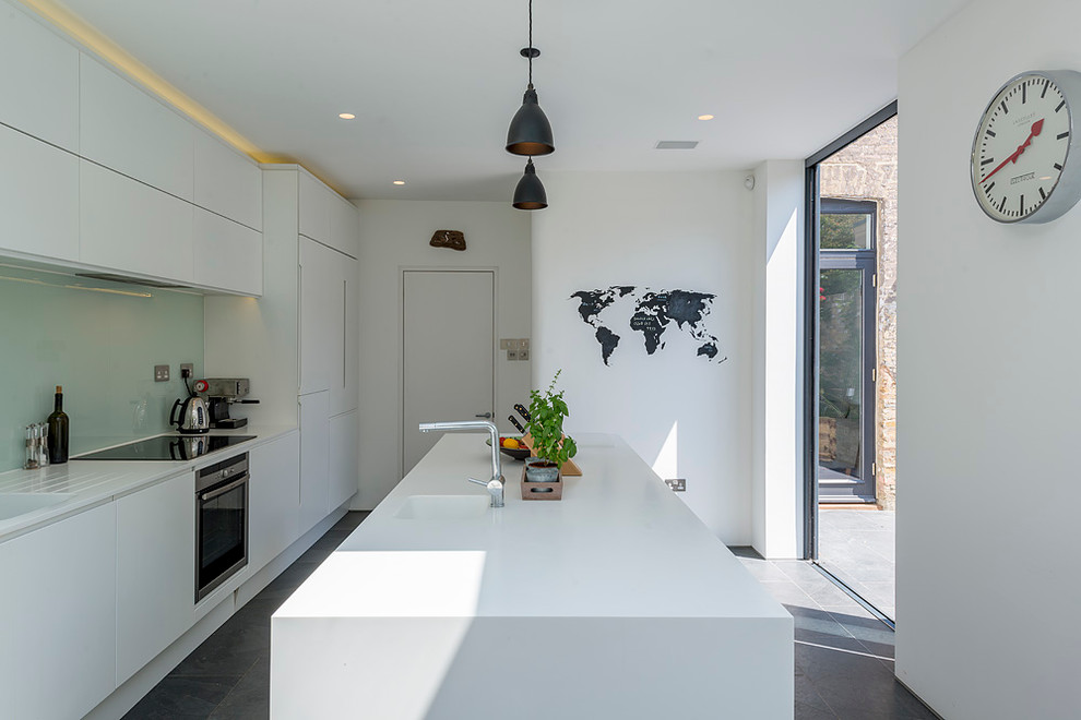 Modern galley enclosed kitchen in London with flat-panel cabinets, white cabinets, blue splashback, glass sheet splashback, white appliances and an island.