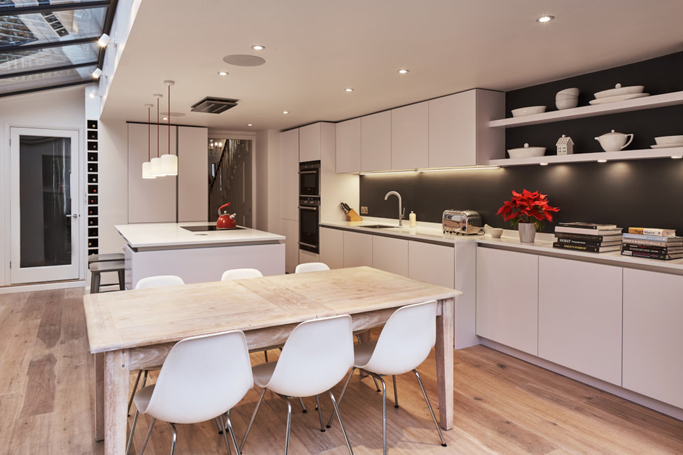 Design ideas for a medium sized contemporary galley kitchen/diner in Kent with an integrated sink, flat-panel cabinets, white cabinets, composite countertops, grey splashback, glass sheet splashback, stainless steel appliances, light hardwood flooring and an island.