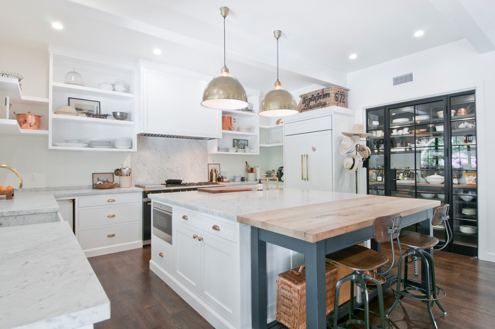 This is an example of a large traditional u-shaped kitchen/diner in Los Angeles with a belfast sink, recessed-panel cabinets, white cabinets, marble worktops, white splashback, marble splashback, black appliances, dark hardwood flooring, an island, brown floors and white worktops.