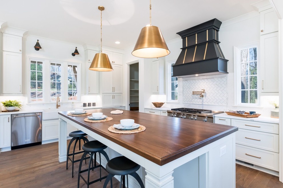This is an example of a classic l-shaped kitchen in Charlotte with a belfast sink, shaker cabinets, white cabinets, white splashback, stainless steel appliances, dark hardwood flooring, an island, brown floors and white worktops.