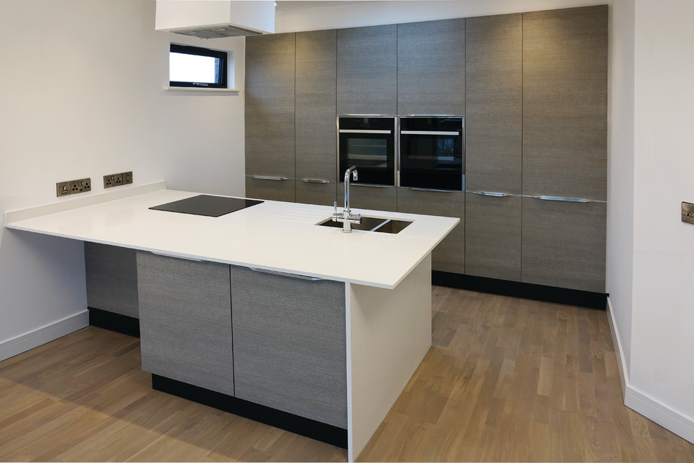 Medium sized modern galley open plan kitchen in Kent with an integrated sink, flat-panel cabinets, light wood cabinets, quartz worktops, black appliances, light hardwood flooring, an island and white worktops.
