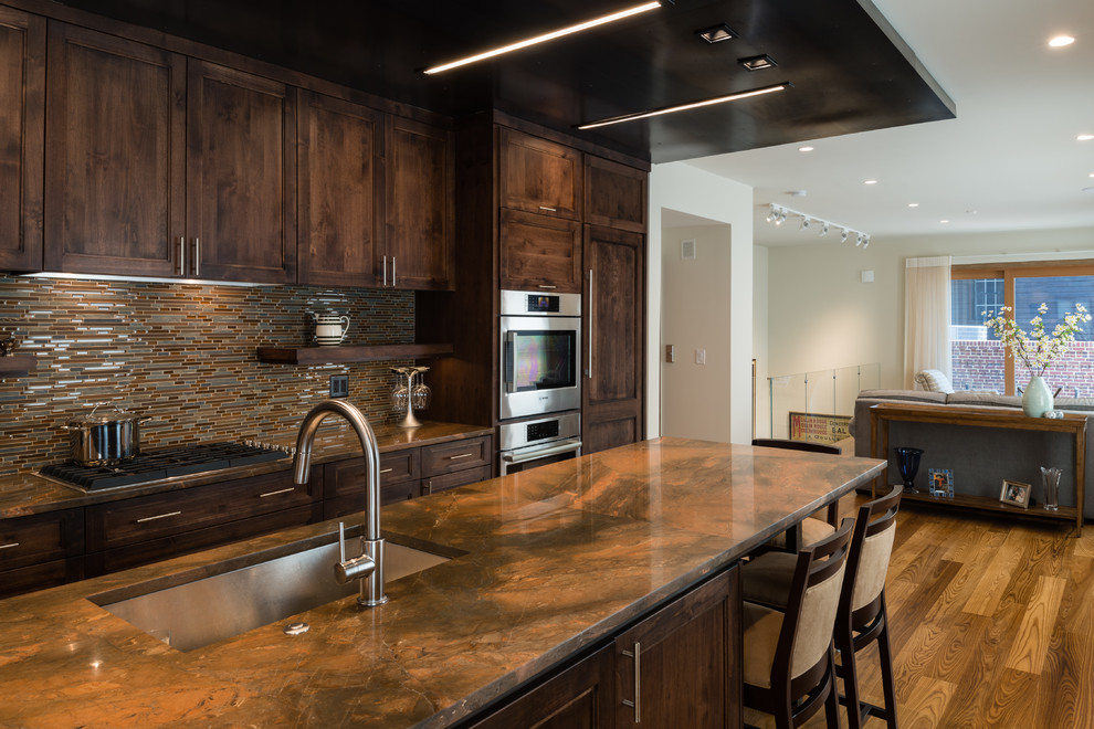 Modern inredning av ett mellanstort kök, med mellanmörkt trägolv, brunt golv, en undermonterad diskho, släta luckor, skåp i mörkt trä, granitbänkskiva, brunt stänkskydd, stänkskydd i glaskakel, integrerade vitvaror och en köksö