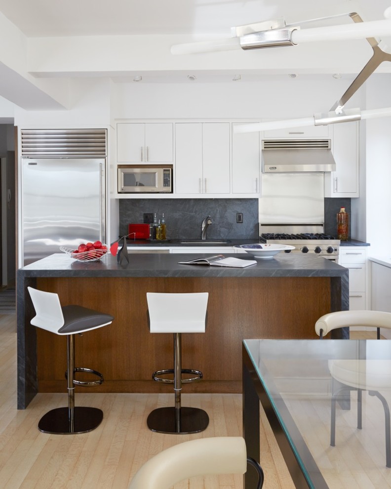 Modern inredning av ett litet linjärt kök och matrum, med en undermonterad diskho, släta luckor, vita skåp, grått stänkskydd, rostfria vitvaror, ljust trägolv, en köksö och stänkskydd i sten