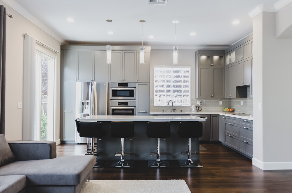 Inspiration for a medium sized contemporary l-shaped open plan kitchen in Sacramento with a double-bowl sink, shaker cabinets, grey cabinets, engineered stone countertops, grey splashback, stainless steel appliances, dark hardwood flooring, an island, brown floors, white worktops and ceramic splashback.