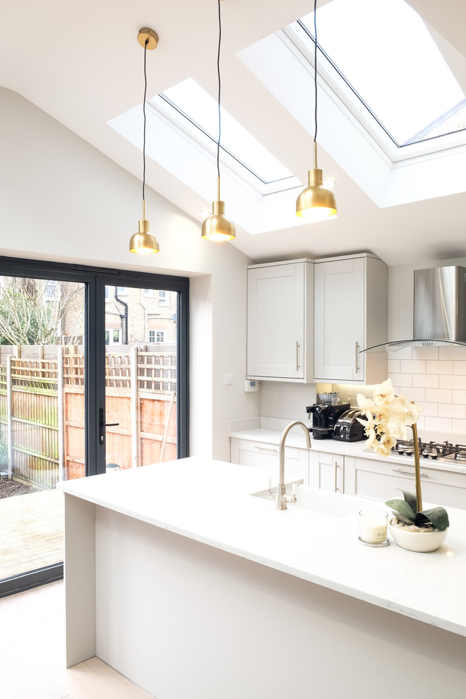 Design ideas for a medium sized classic single-wall open plan kitchen in Sussex with a belfast sink, shaker cabinets, grey cabinets, quartz worktops, white splashback, metro tiled splashback, stainless steel appliances, light hardwood flooring, an island, brown floors and white worktops.