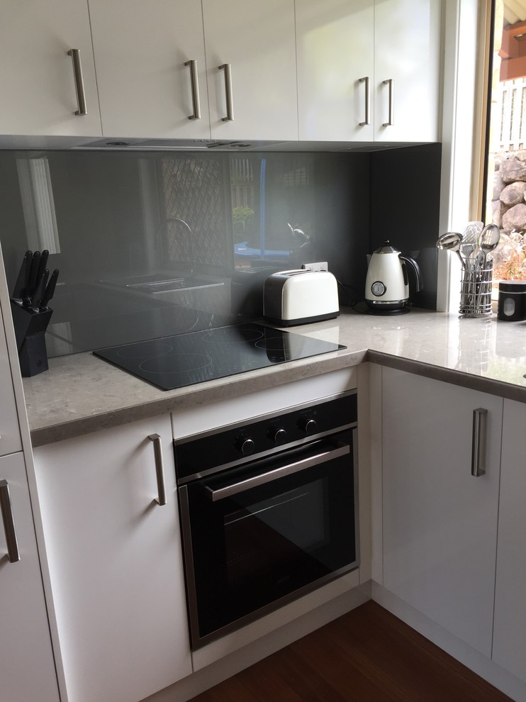 Exempel på ett avskilt, litet modernt u-kök, med en dubbel diskho, vita skåp, laminatbänkskiva, grått stänkskydd, glaspanel som stänkskydd, svarta vitvaror, mörkt trägolv, en halv köksö och brunt golv