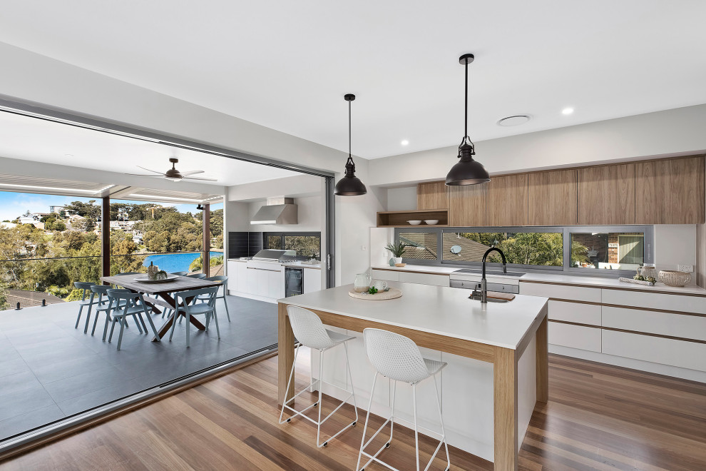 Zweizeilige, Geräumige Moderne Wohnküche mit Einbauwaschbecken, flächenbündigen Schrankfronten, weißen Schränken, Küchengeräten aus Edelstahl, braunem Holzboden, Kücheninsel, braunem Boden und weißer Arbeitsplatte in Wollongong