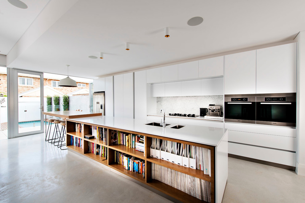 На фото: параллельная кухня в современном стиле с двойной мойкой, плоскими фасадами, белыми фасадами, белым фартуком, техникой из нержавеющей стали, бетонным полом и островом