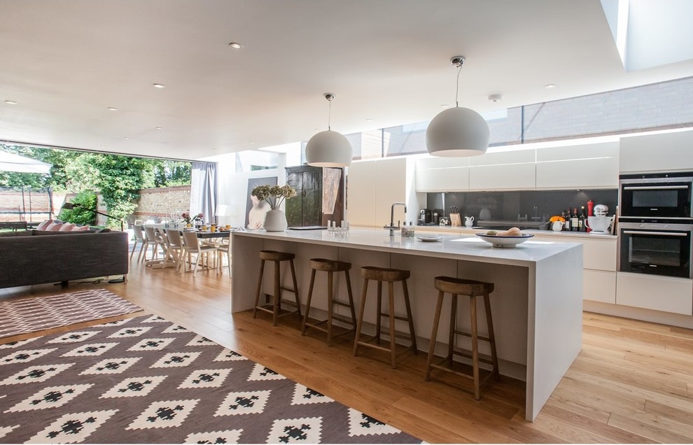 Contemporary kitchen in London with flat-panel cabinets, white cabinets, grey splashback, stainless steel appliances, medium hardwood flooring and an island.