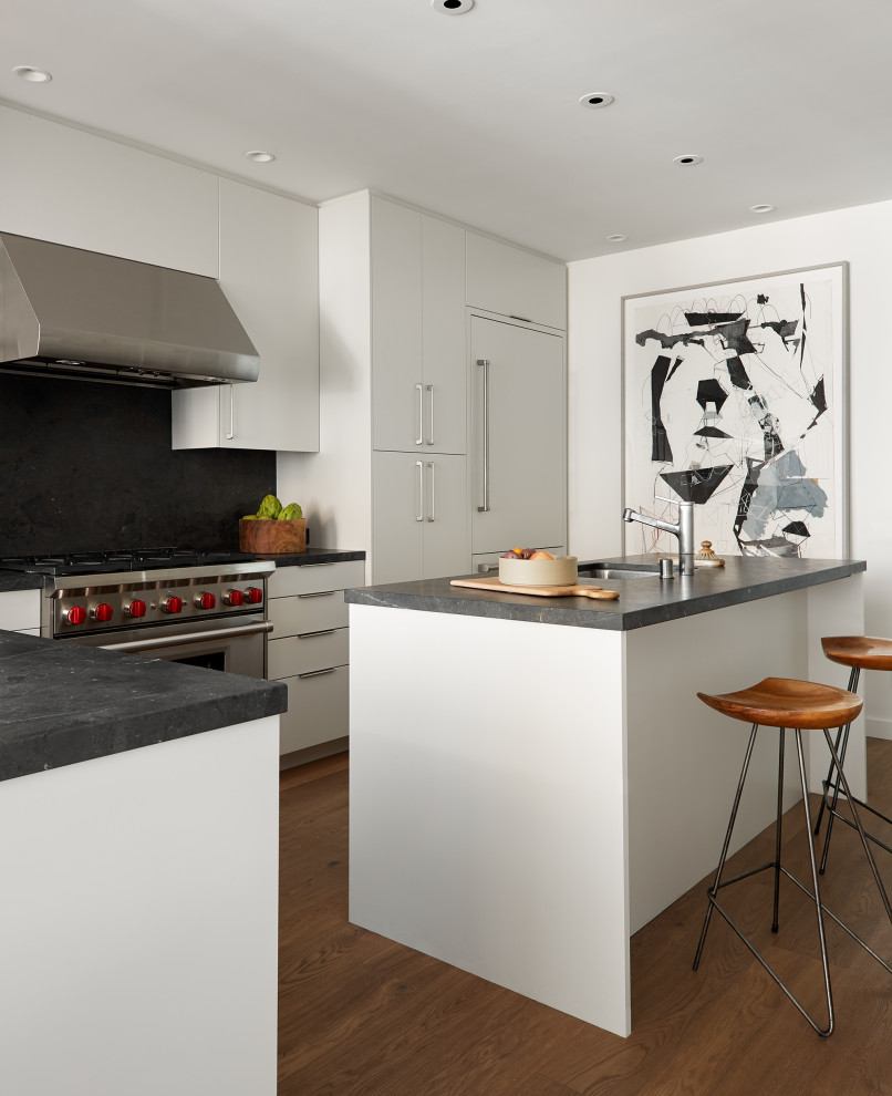 Inspiration for an urban l-shaped kitchen in San Francisco with a submerged sink, flat-panel cabinets, white cabinets, black splashback, stone slab splashback, integrated appliances, medium hardwood flooring, an island, brown floors and black worktops.