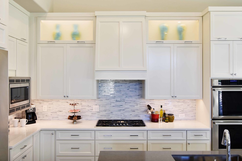Klassisk inredning av ett stort grå grått kök, med en undermonterad diskho, luckor med infälld panel, vita skåp, bänkskiva i kvarts, vitt stänkskydd, stänkskydd i stenkakel, rostfria vitvaror, klinkergolv i porslin, en köksö och beiget golv