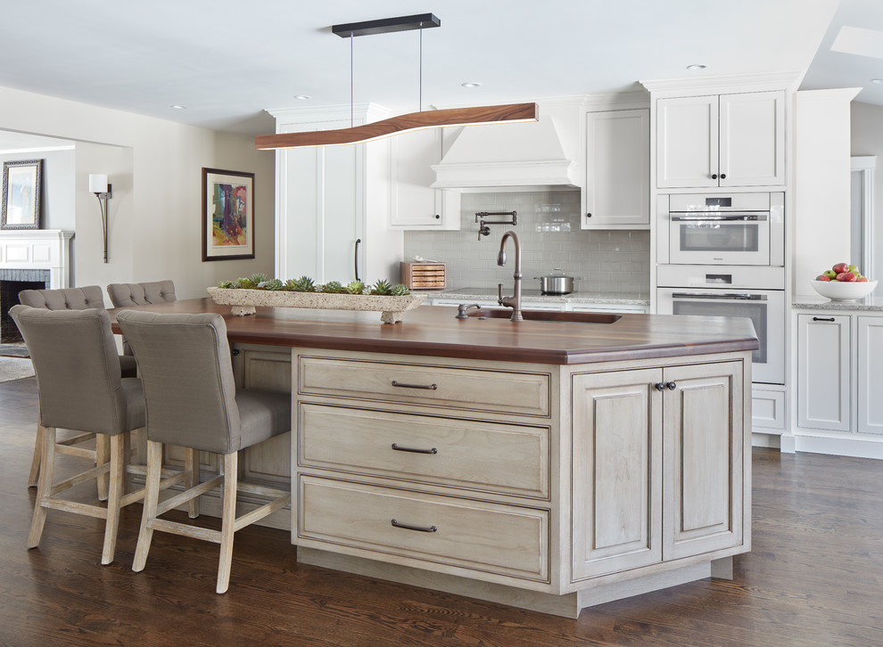 Mid-sized transitional l-shaped dark wood floor and brown floor open concept kitchen photo in Boston with a single-bowl sink, wood countertops, gray backsplash, glass tile backsplash, white appliances, an island, raised-panel cabinets, distressed cabinets and gray countertops