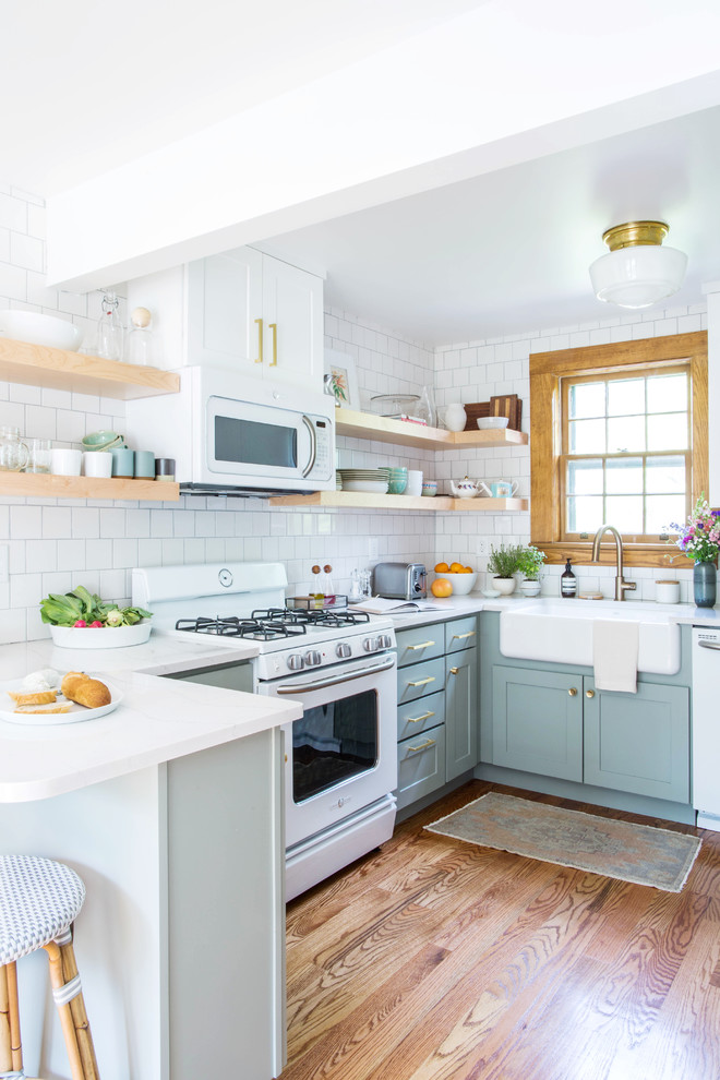 Example of a small transitional u-shaped medium tone wood floor and brown floor eat-in kitchen design in Other with a farmhouse sink, shaker cabinets, blue cabinets, quartz countertops, white backsplash, porcelain backsplash, white appliances and a peninsula
