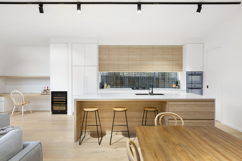 Design ideas for a contemporary galley open plan kitchen in Melbourne with a submerged sink, flat-panel cabinets, white cabinets, window splashback, integrated appliances, light hardwood flooring, an island, beige floors and white worktops.