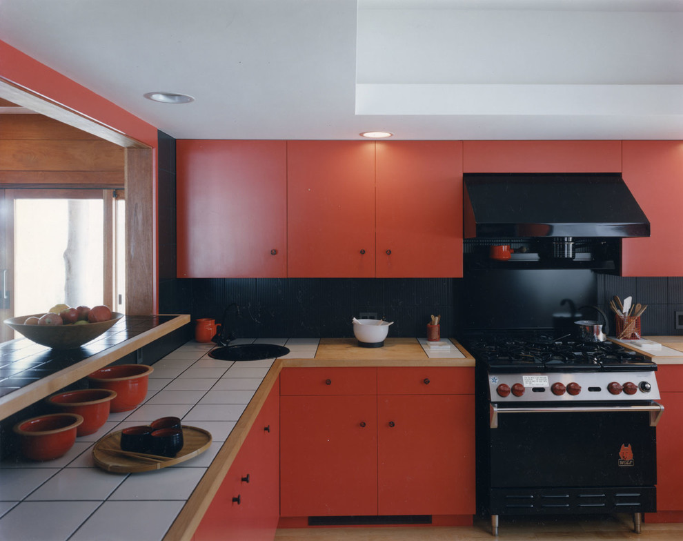 Modern inredning av ett avskilt, mellanstort l-kök, med en nedsänkt diskho, släta luckor, röda skåp, kaklad bänkskiva, svart stänkskydd, stänkskydd i porslinskakel, svarta vitvaror och ljust trägolv