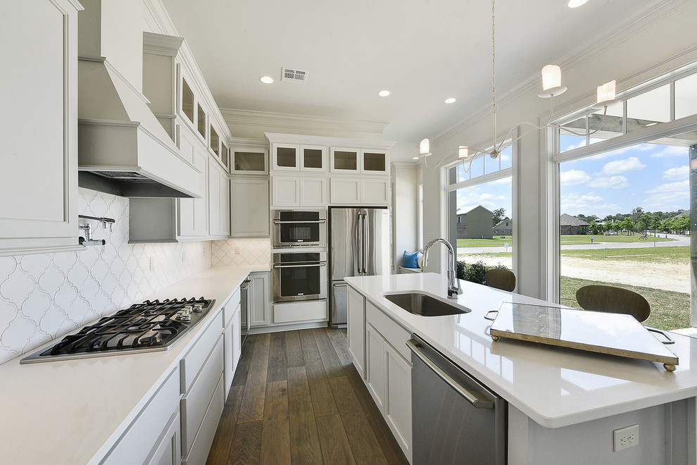 Example of a mid-sized trendy l-shaped medium tone wood floor eat-in kitchen design in New Orleans with an island, a drop-in sink, shaker cabinets, white cabinets, quartz countertops, white backsplash, glass tile backsplash and stainless steel appliances