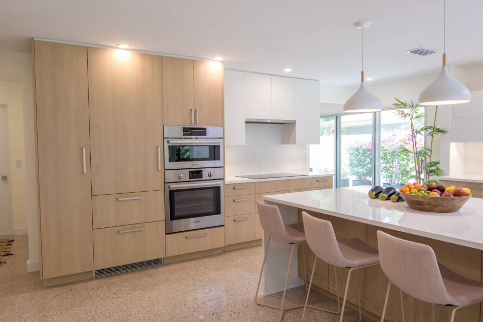 Inspiration for a large modern l-shaped open plan kitchen in Tampa with a belfast sink, flat-panel cabinets, light wood cabinets, composite countertops, white splashback, stainless steel appliances, terrazzo flooring, an island, multi-coloured floors and white worktops.
