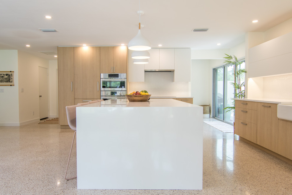 Large modern l-shaped open plan kitchen in Tampa with a belfast sink, flat-panel cabinets, light wood cabinets, composite countertops, white splashback, stainless steel appliances, terrazzo flooring, an island, multi-coloured floors and white worktops.