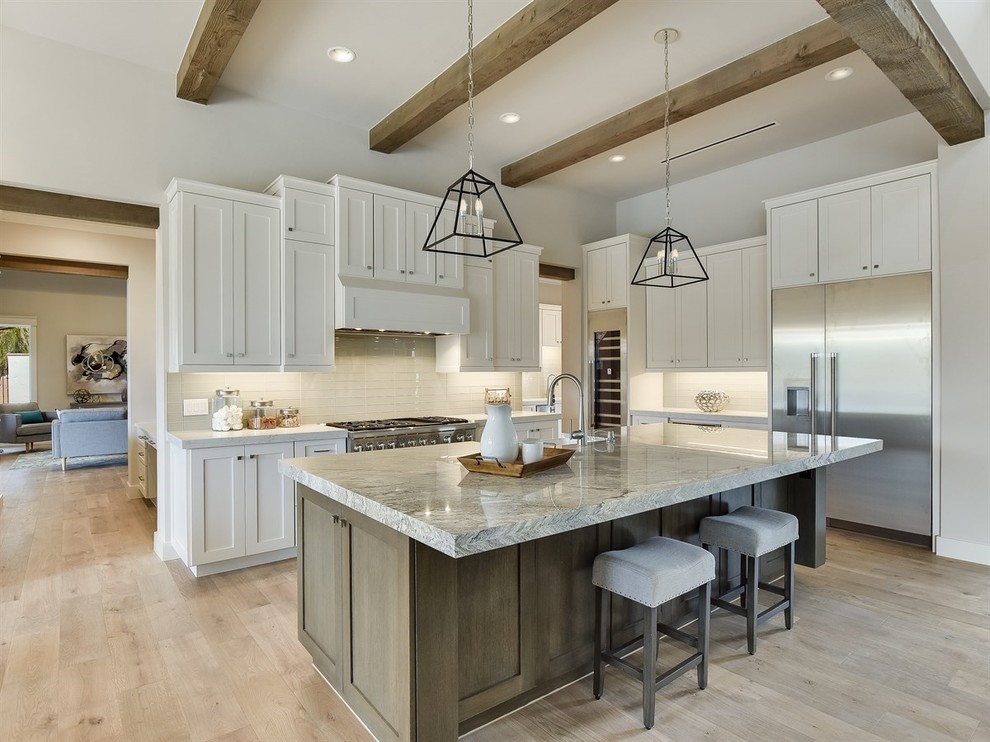This is an example of a traditional l-shaped kitchen in Austin with shaker cabinets, white cabinets, white splashback, stainless steel appliances, light hardwood flooring, an island, beige floors and grey worktops.