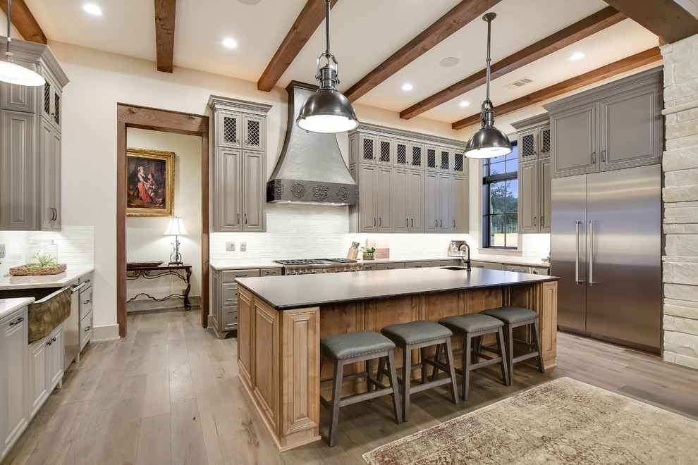 Design ideas for a country u-shaped kitchen in Austin with a belfast sink, raised-panel cabinets, grey cabinets, white splashback, stainless steel appliances, medium hardwood flooring, an island and brown floors.