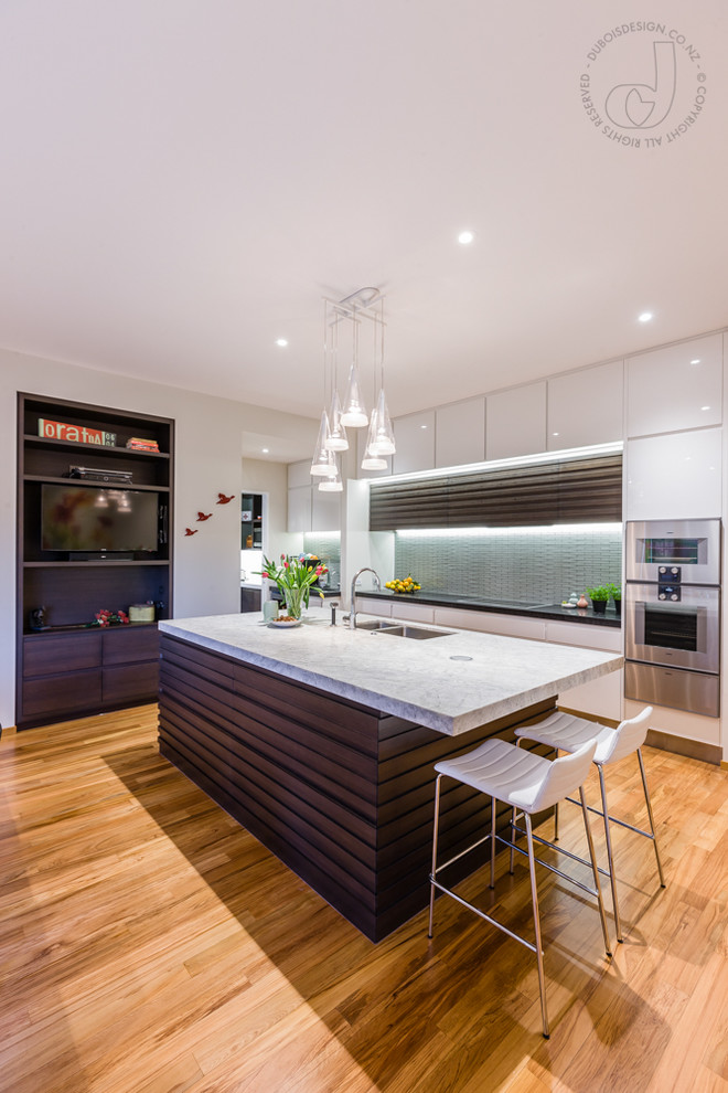 Photo of a medium sized contemporary galley kitchen/diner in Auckland with a double-bowl sink, louvered cabinets, dark wood cabinets, quartz worktops, green splashback, glass tiled splashback, stainless steel appliances, light hardwood flooring and an island.
