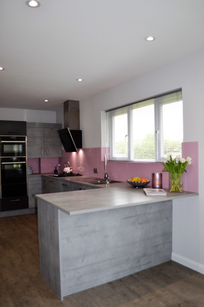 This is an example of a medium sized contemporary u-shaped kitchen/diner in Other with a built-in sink, flat-panel cabinets, grey cabinets, laminate countertops, pink splashback, glass sheet splashback, black appliances and a breakfast bar.
