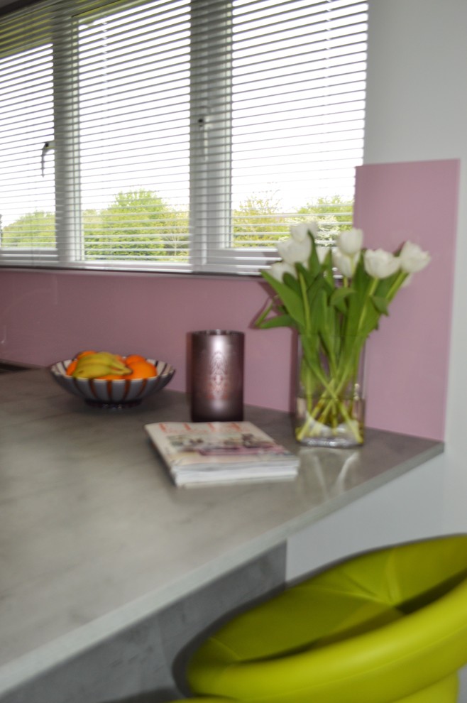 This is an example of a medium sized contemporary u-shaped kitchen/diner in Other with a built-in sink, flat-panel cabinets, grey cabinets, laminate countertops, pink splashback, glass sheet splashback, black appliances and a breakfast bar.
