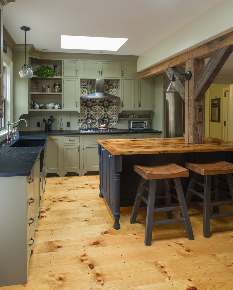 Foto på ett stort lantligt l-kök, med en rustik diskho, luckor med profilerade fronter, gröna skåp, bänkskiva i täljsten, brunt stänkskydd, stänkskydd i keramik, rostfria vitvaror, ljust trägolv och en köksö
