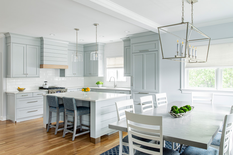 Nautical l-shaped kitchen pantry in New York with a submerged sink, shaker cabinets, blue cabinets, engineered stone countertops, white splashback, metro tiled splashback, integrated appliances, light hardwood flooring, an island and white worktops.