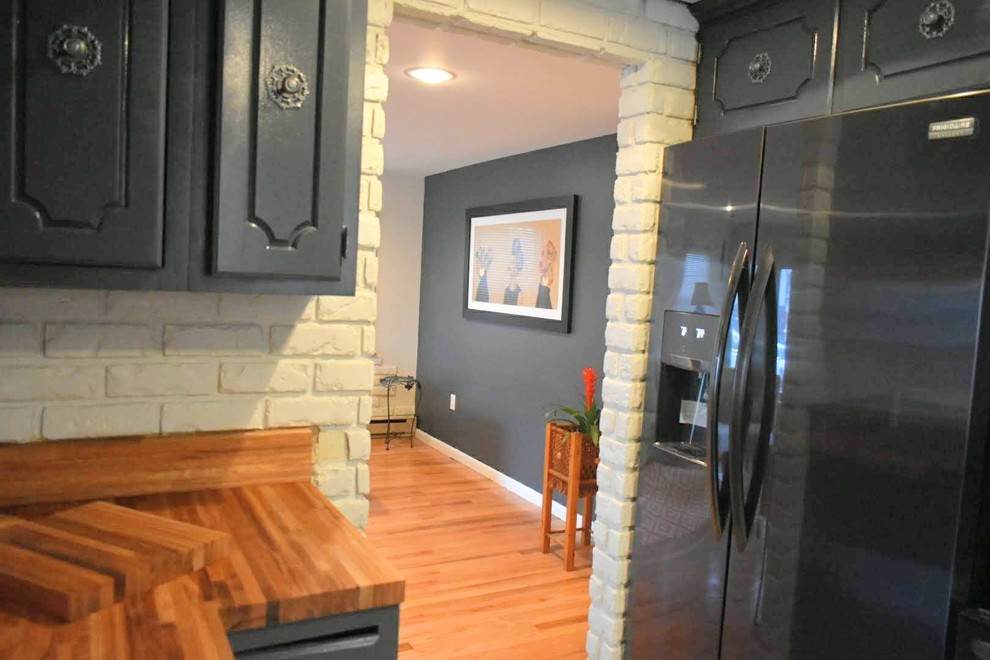 Foto på ett retro kök, med blå skåp, träbänkskiva, stänkskydd i tegel, svarta vitvaror och ljust trägolv