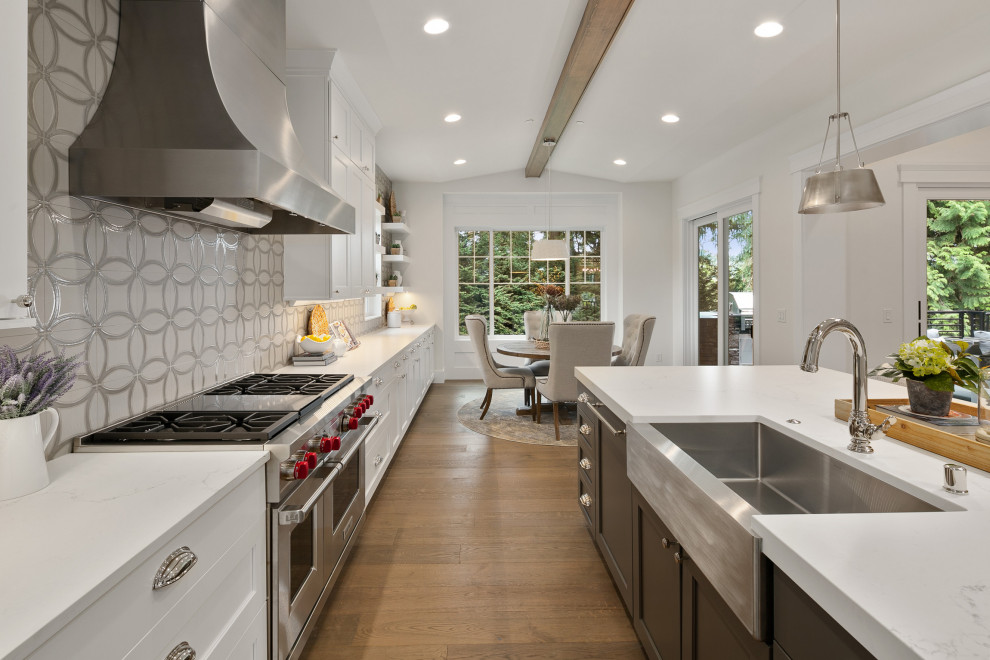 Photo of a large classic single-wall kitchen/diner in Seattle with a belfast sink, shaker cabinets, white cabinets, quartz worktops, grey splashback, porcelain splashback, stainless steel appliances, medium hardwood flooring, an island, brown floors, white worktops and exposed beams.