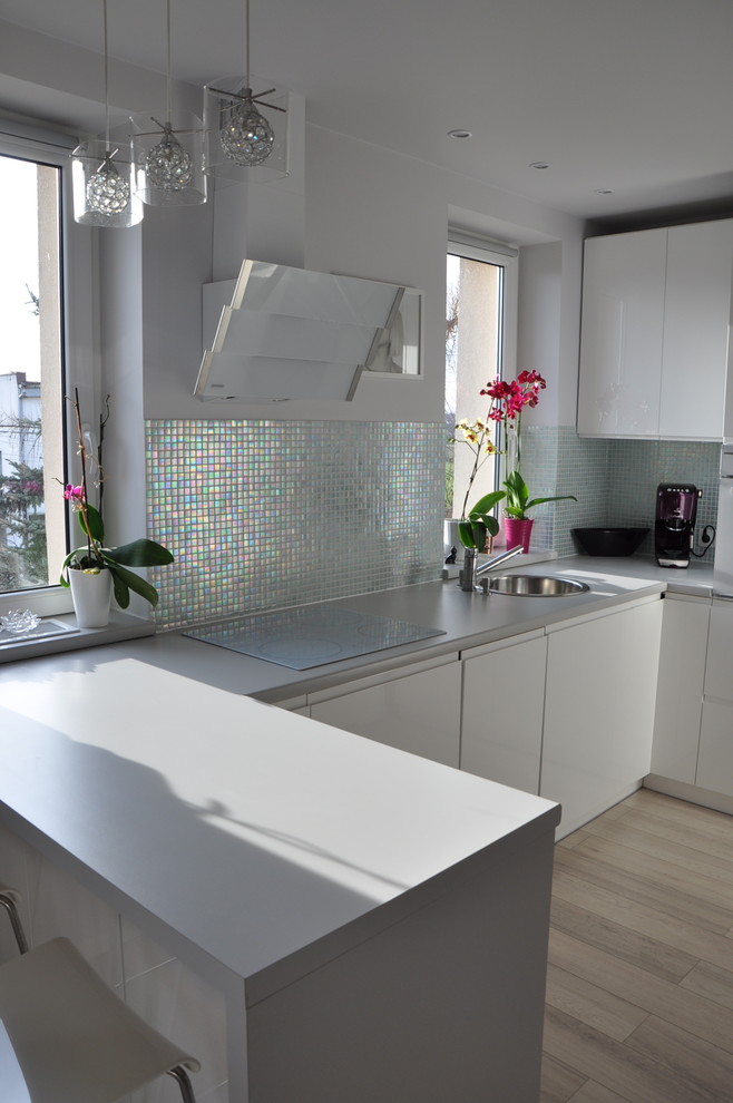 This is an example of a small contemporary u-shaped enclosed kitchen in Other with a single-bowl sink, flat-panel cabinets, white cabinets, laminate countertops, metallic splashback, mosaic tiled splashback, white appliances, light hardwood flooring and an island.