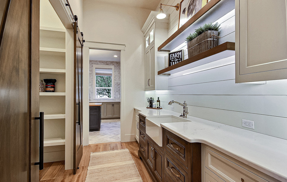 Design ideas for an expansive country u-shaped kitchen pantry in Portland with a belfast sink, recessed-panel cabinets, white cabinets, engineered stone countertops, white splashback, ceramic splashback, stainless steel appliances, light hardwood flooring, an island, brown floors and white worktops.