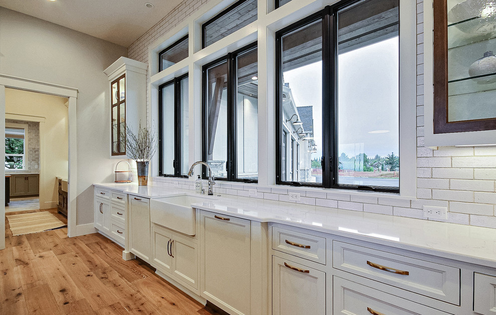 Bild på ett mycket stort lantligt vit vitt kök, med en rustik diskho, luckor med infälld panel, vita skåp, bänkskiva i kvarts, vitt stänkskydd, stänkskydd i keramik, rostfria vitvaror, ljust trägolv, en köksö och brunt golv