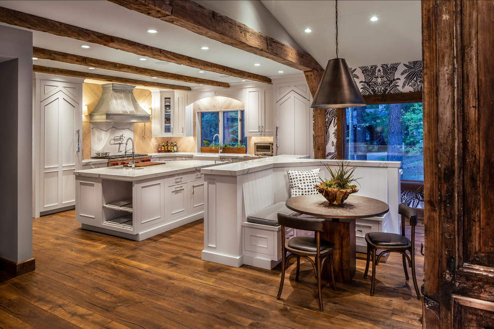 Rustic l-shaped kitchen/diner in Other with a belfast sink, recessed-panel cabinets, white cabinets, white splashback, stone slab splashback, stainless steel appliances, multiple islands, white worktops, quartz worktops, dark hardwood flooring and brown floors.