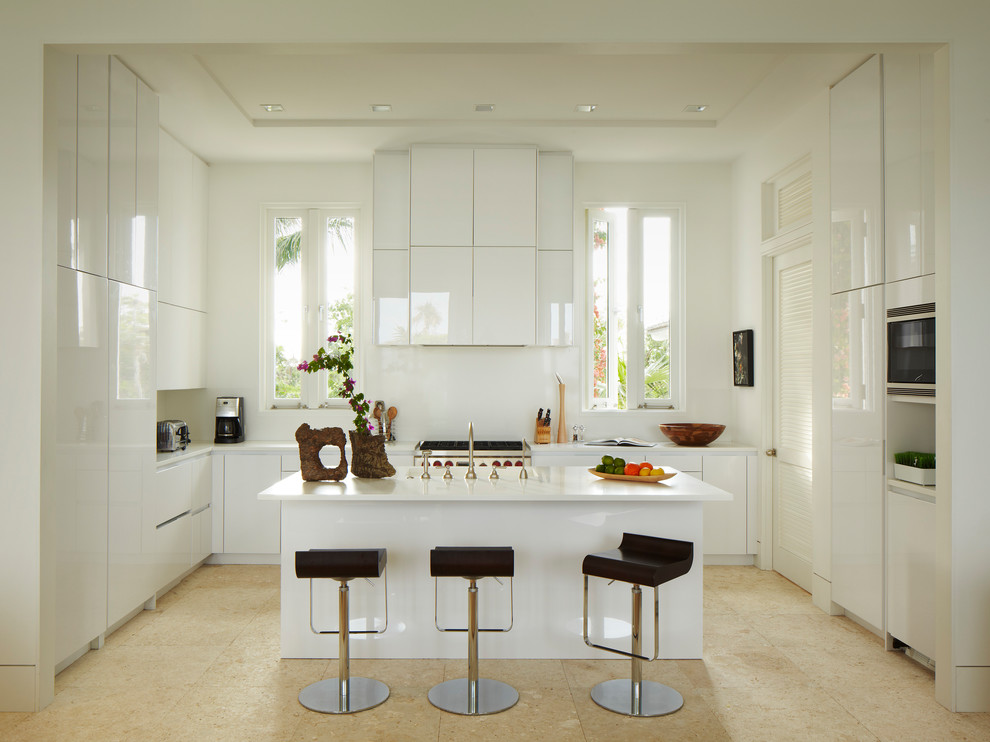 На фото: кухня в морском стиле с плоскими фасадами, белыми фасадами, белым фартуком, техникой из нержавеющей стали, островом и бежевым полом