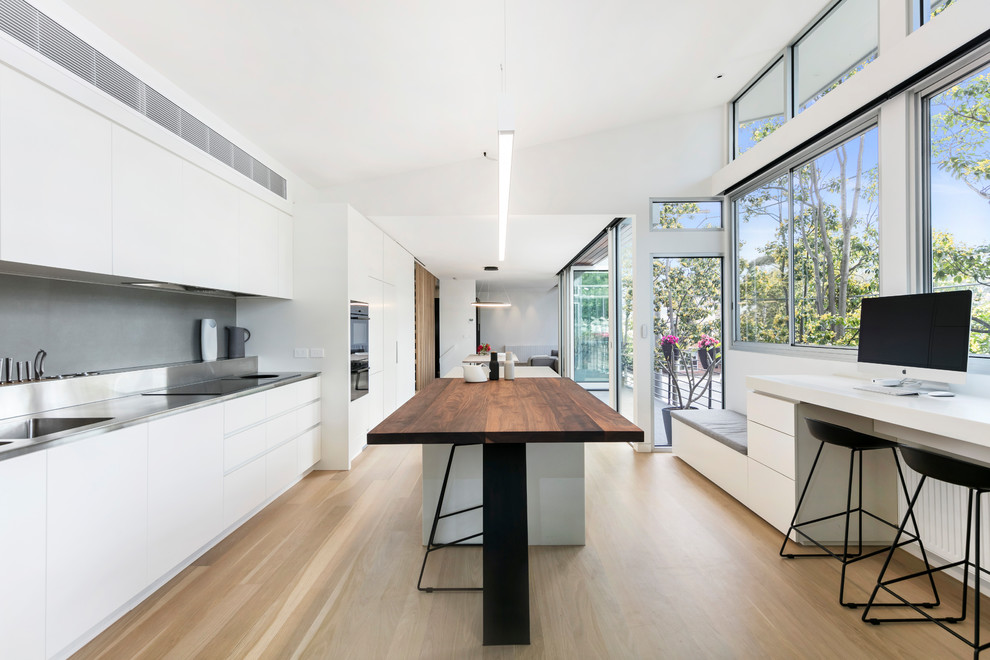 Inredning av ett modernt kök, med en integrerad diskho, släta luckor, vita skåp, bänkskiva i rostfritt stål, grått stänkskydd, integrerade vitvaror, ljust trägolv, en köksö och beiget golv