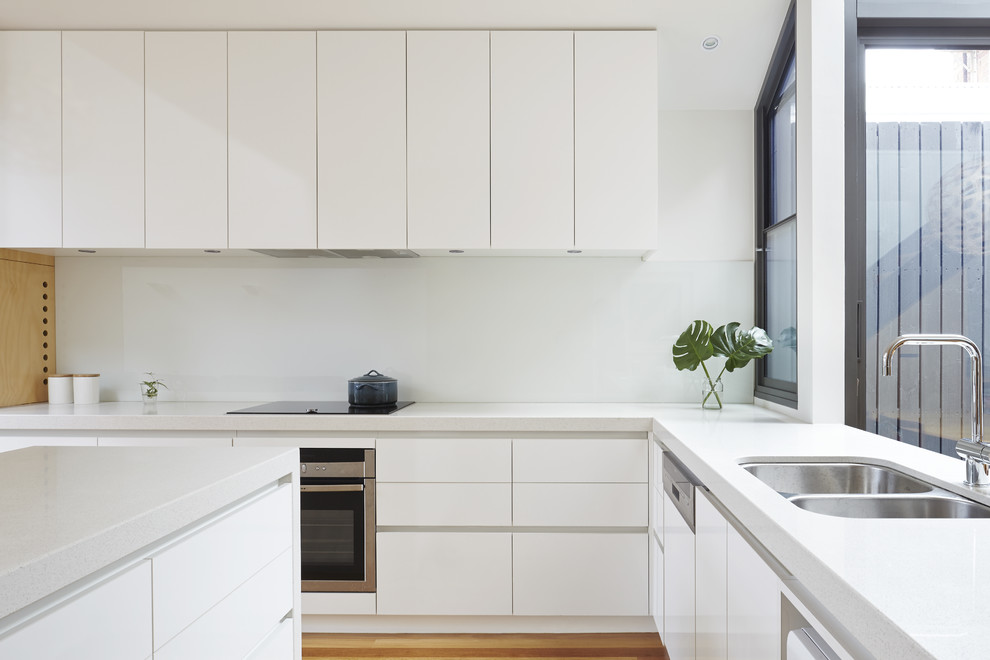Design ideas for a medium sized contemporary l-shaped kitchen/diner in Melbourne with a submerged sink, flat-panel cabinets, white cabinets, engineered stone countertops, white splashback, glass sheet splashback, stainless steel appliances, medium hardwood flooring and an island.