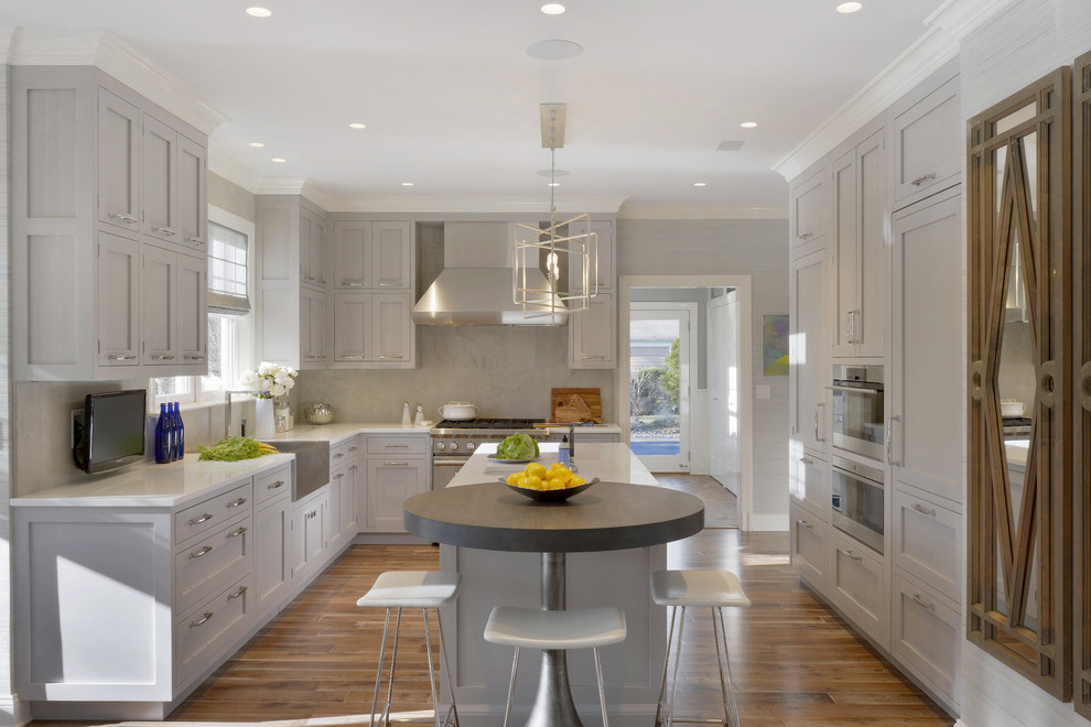 Example of a huge classic u-shaped medium tone wood floor and brown floor eat-in kitchen design in New York with a farmhouse sink, shaker cabinets, gray cabinets, glass countertops, gray backsplash, limestone backsplash, paneled appliances and an island