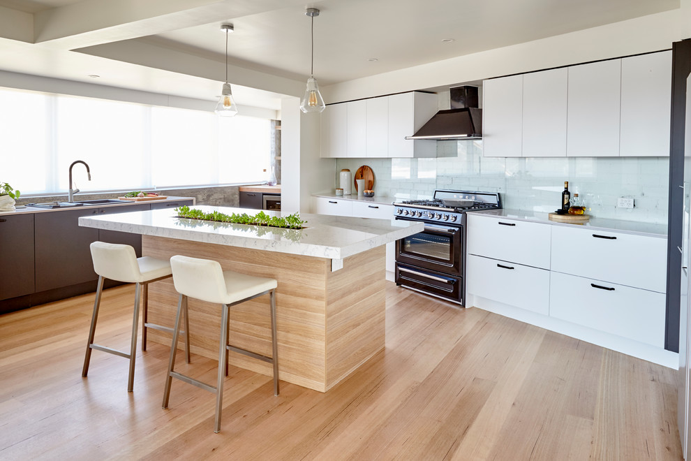 Große Moderne Küche mit flächenbündigen Schrankfronten, Marmor-Arbeitsplatte, hellem Holzboden, Kücheninsel, Einbauwaschbecken, weißen Schränken, Küchenrückwand in Weiß und schwarzen Elektrogeräten in Melbourne