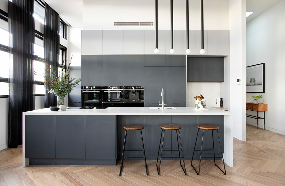 This is an example of a contemporary galley kitchen in Sydney with a submerged sink, flat-panel cabinets, grey cabinets, black appliances, medium hardwood flooring, an island, brown floors and white worktops.