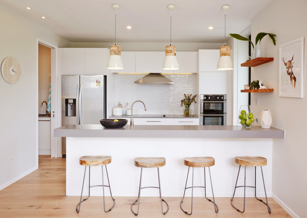 Design ideas for a scandinavian galley kitchen in Auckland with flat-panel cabinets, white cabinets, white splashback, stainless steel appliances, light hardwood flooring and a breakfast bar.