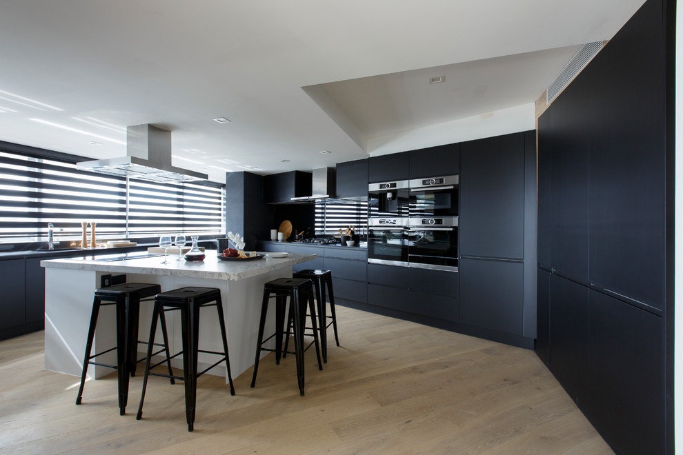 Cette image montre une grande cuisine design avec un placard à porte plane, plan de travail en marbre, une crédence noire, îlot, un électroménager noir et parquet clair.
