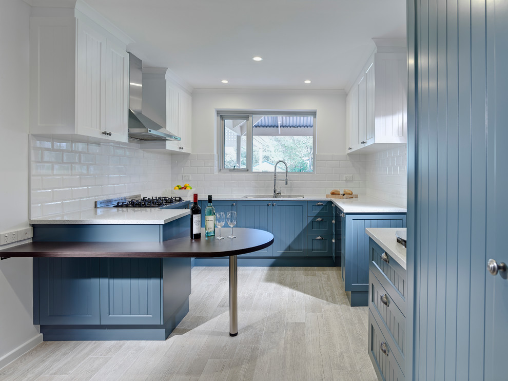Klassisk inredning av ett avskilt, litet u-kök, med en undermonterad diskho, luckor med infälld panel, blå skåp, vitt stänkskydd, stänkskydd i tunnelbanekakel, rostfria vitvaror, en halv köksö och beiget golv