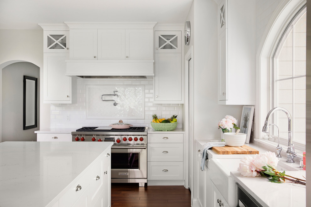 Cette image montre une cuisine ouverte traditionnelle en L de taille moyenne avec un évier de ferme, un placard à porte shaker, des portes de placard blanches, un plan de travail en quartz modifié, une crédence blanche, une crédence en céramique, un électroménager en acier inoxydable, un sol en bois brun, îlot, un sol marron et un plan de travail blanc.