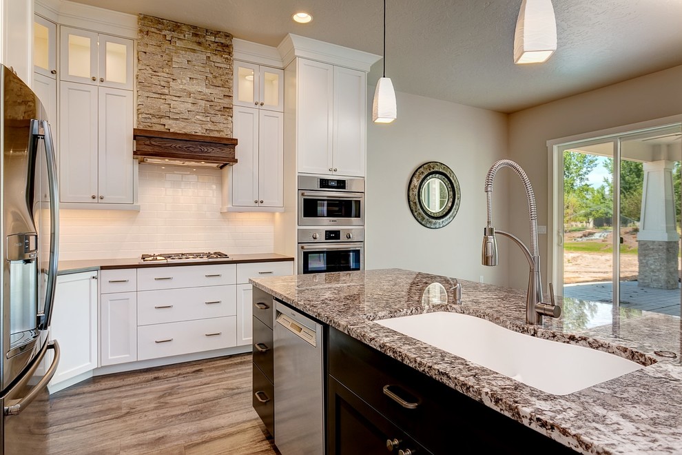Example of a large arts and crafts l-shaped medium tone wood floor open concept kitchen design in Boise with an undermount sink, shaker cabinets, white cabinets, granite countertops, white backsplash, porcelain backsplash, stainless steel appliances and an island