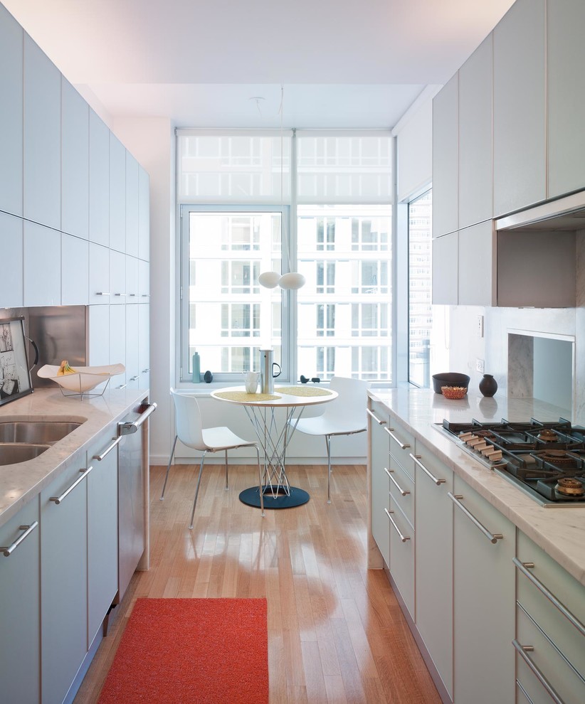 Cette image montre une cuisine américaine parallèle design avec un évier encastré, un placard à porte plane, des portes de placard blanches, un électroménager en acier inoxydable et un sol en bois brun.