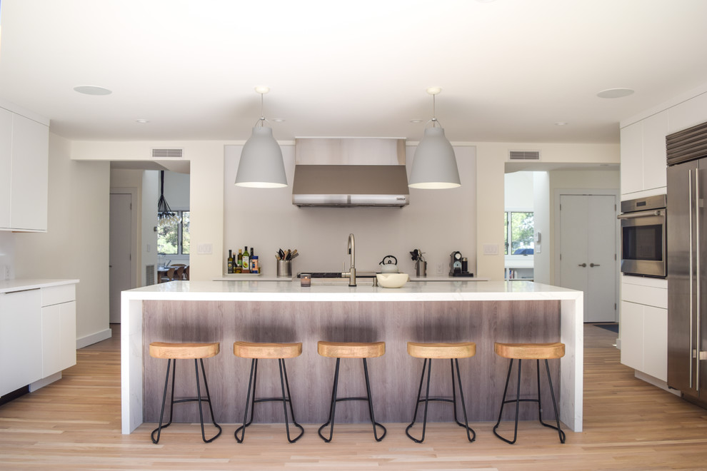 Large contemporary u-shaped kitchen in New York with a submerged sink, flat-panel cabinets, white cabinets, engineered stone countertops, white splashback, stone slab splashback, stainless steel appliances, light hardwood flooring, an island, white worktops and beige floors.